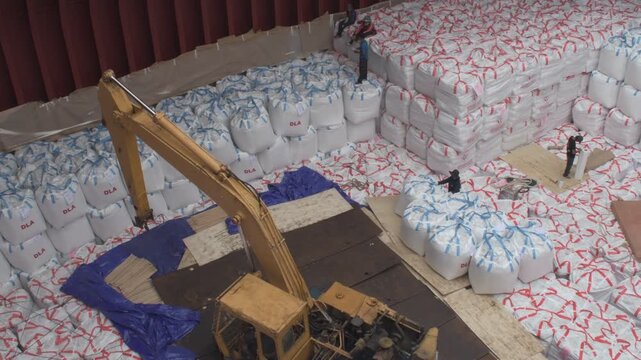 high angle view inside a bulk carrier cargo hold showing a yellow excavator and stevedores neatly stowing tiers of white jumbo cargo bags during loading operations