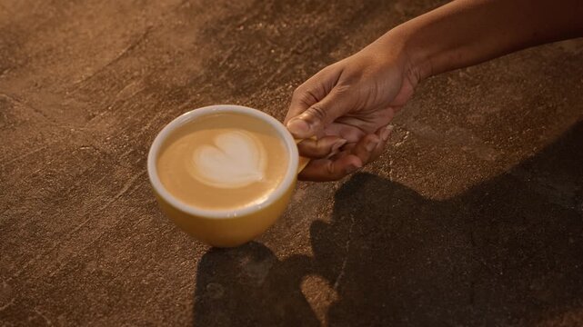 Hand placing yellow cup with heart latte art on table in slow motion, top view. Video suitable for cafe branding, menu marketing and coffee shop ads