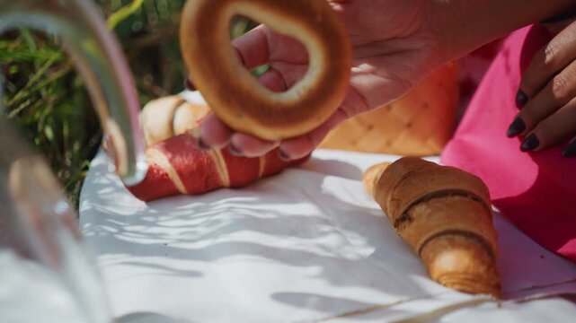 Close up of woman hand reaching for and picking up bagels from outdoor picnic setup with croissants bread basket on white cloth under sunlight. scene includes soft shadows cast by foliage