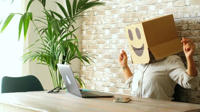 Creative businesswoman wearing a smiley face cardboard box on her head, dancing joyfully at her office desk with a laptop, celebrating a successful business achievement and happiness at work