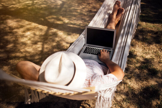 Man relaxing in a hammock using a laptop computer outdoors, wearing a white hat, with bare feet visible on a sunny day in a natural setting