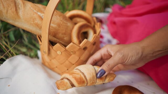 Close up of hand reaching for and picking up croissants from woven basket on white cloth with grass in background. Hand with purple nail polish interacts with bread items including bagels