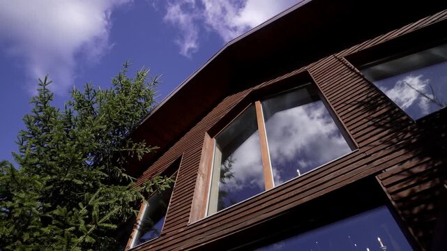 Low-angle pan over wooden house facade with reflective windows and adjacent evergreen under blue sky, with house, facade, wood, siding detail, showing window, glass, reflection, roof context