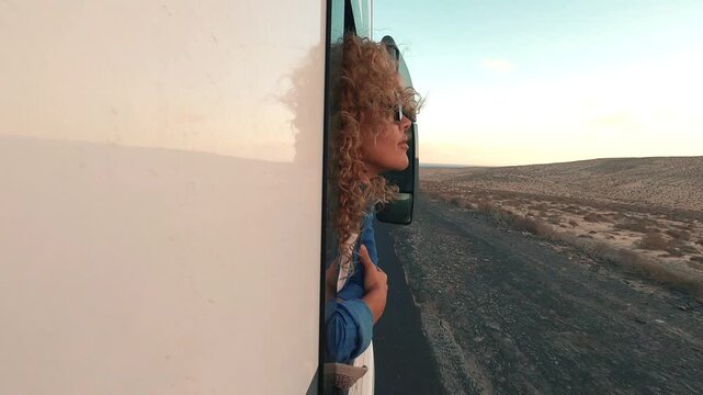 Joyful woman with curly hair feeling the wind while looking out the window of a moving camper van, enjoying the scenic desert landscape during an unforgettable summer vacation road trip