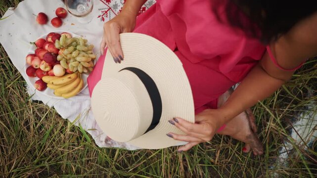 Close up of woman in pink adjusting her straw hat while sitting on grass with fruit and wine. white cloth grapes apples bananas red berries glassware are visible around her. Soft natural