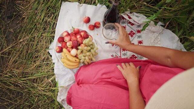 Woman in pink dress enjoys outdoor picnic on grass reaching for wine and interacting with fruit. white cloth holds apples bananas grapes loose cherries dark bottle of drink glassware red