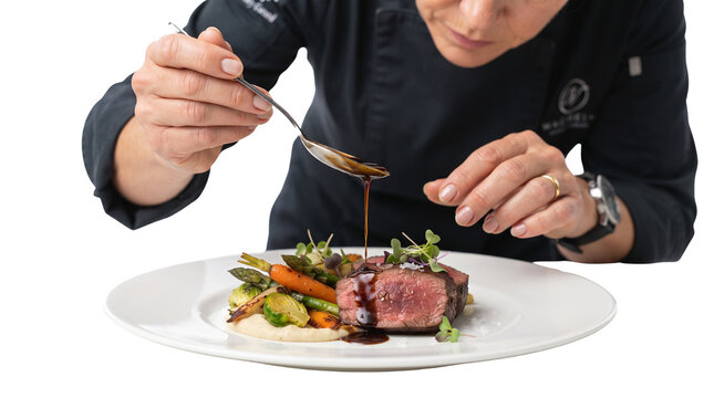 Professional chef plating gourmet beef steak, culinary expert drizzling sauce on meat dish, fine dining food preparation on white background