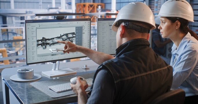 Female Specialist Points to CAD Graphics on Screen as Male Engineer Checks Machined Part with Caliper. Quality Assurance and Digital Twin Technology Optimize Smart Factory Automotive Workflows.
