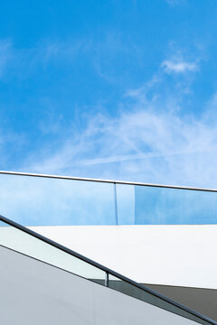 Minimal white rooftop terrace with glass railing and diagonal lines sits beneath blue sky in a view where the terrace and railing define the entire frame