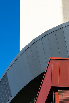 Curved gray and red architectural forms meet a white facade beneath blue sky in an abstract view where the architectural forms overlap across the frame