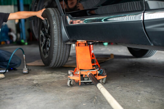Selective focus to hydraulic floor jack with blurry hand of professional mechanic changing a car tire lifted and red jack stand inside an auto repair shop. Focus on equipment and car maintenance.