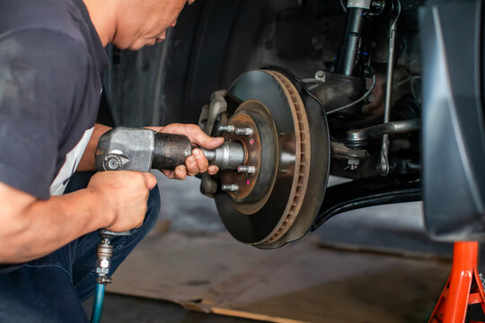 Selective focus to professional mechanic using a pneumatic impact wrench to service the brake disc assembly and wheel hub of a car supported by a jack stand in a repair shop.