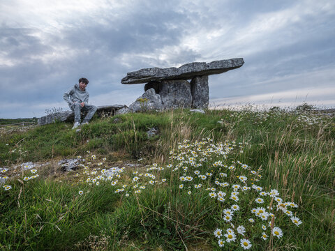 Young explorer sits near ancient stone monument in Poulnabrone. Daisies bloom wild across green grassy field. Sky clouds loom over historic structure. Child gazes upward with curious wonder
