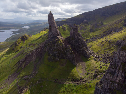 Aerial view of the Old Man of Storr rock formation on the Isle of Skye, featuring dramatic basalt pinnacles, lush green grassy slopes, and a distant lake in Scotland.
