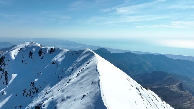 Aerial view of Pania della Croce peak covered in deep snow under a bright blue sky with the Mediterranean Sea visible in the distance in Stazzema, Tuscany, Italy.