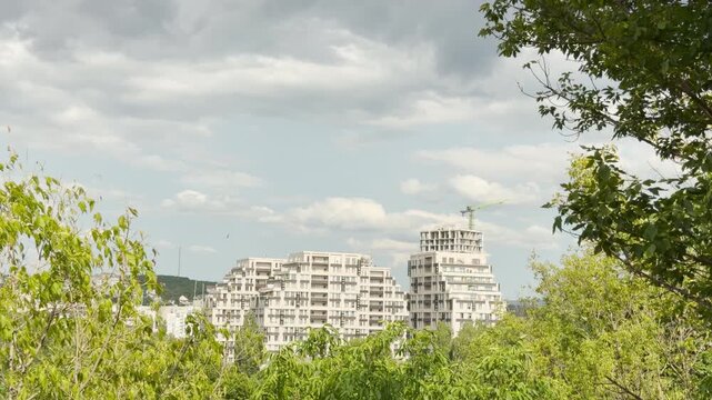 Modern high-rise buildings rise beyond leafy green trees in Simferopol, Crimea. A construction crane stands against the cloudy sky. The bright urban scene brings a hopeful sense of calm.