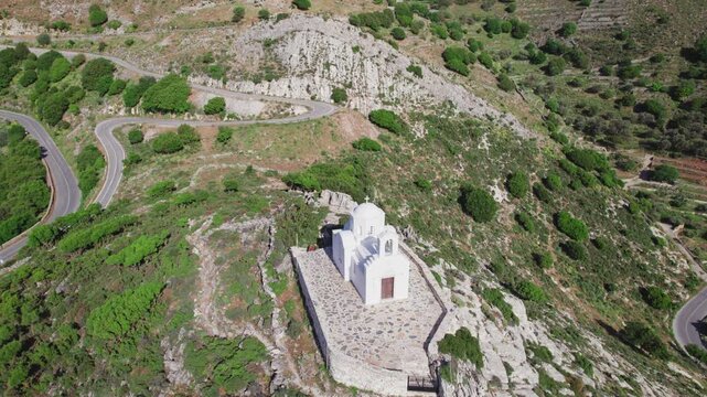 The drone ascends over a pristine whitewashed chapel on a Naxos ridge, revealing winding mountain roads and rugged green hills framing the lone Greek sanctuary