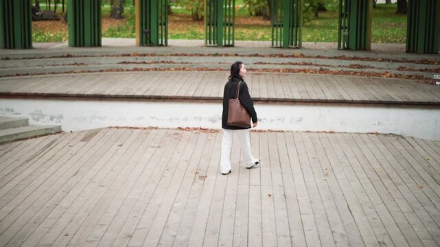 Young woman in black coat and white pants walks slowly across empty wooden deck carrying brown bag over her shoulder. scene features green structural columns with autumn leaves scattered