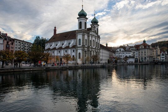 Lucerne, Switzerland - 01 November 2018: View of the Jesuit Church reflecting in the Reuss River with historic buildings and a cloudy sky in the background.