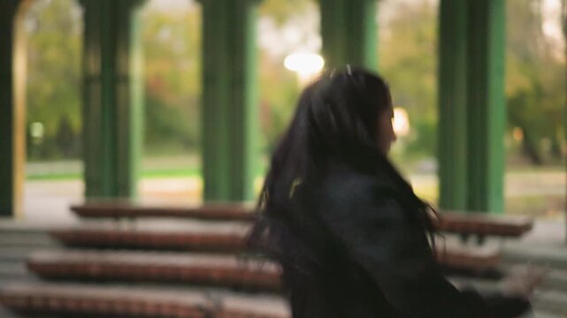 Close up of young woman dancing gracefully in open pavilion with green pillars and blurred park background. Her long dark hair flows freely as woman moves wearing black blazer over white top