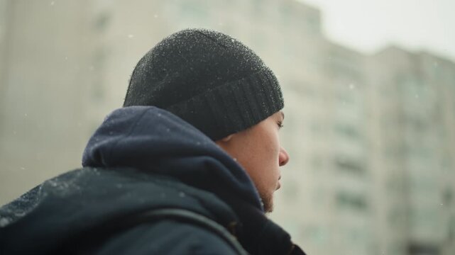 Close up of man wearing winter clothing including black knit hat and dark blue jacket standing outdoors in falling snow. subject remains mostly stationary with minimal movement as light gray