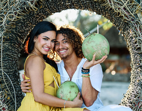 A smiling couple shares fresh coconuts while relaxing inside a woven beach nest on a sunlit tropical shore. The woman in a yellow dress and her partner laugh together, capturing a carefree summer vaca