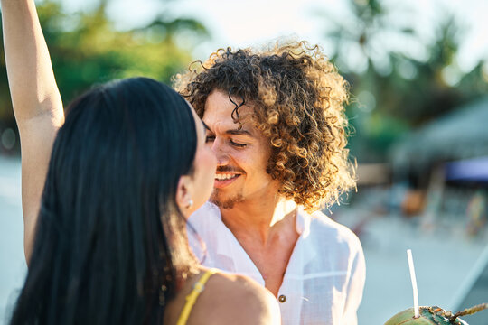 A young couple laugh and dance along a tropical beach, the man holding a fresh coconut as waves lap the shoreline. The scene captures romantic summer vacation vibes, carefree joy and togetherness amid