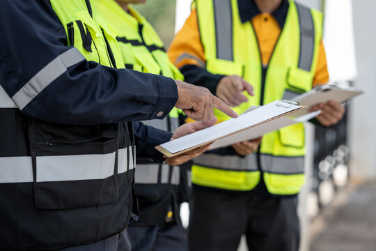 Construction workers in high visibility safety vests review documents on a clipboard during an on-site inspection and project meeting.