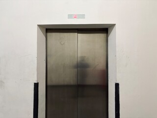Closed stainless steel elevator doors in a minimalist white interior, featuring a digital floor indicator above the entrance. The clean architectural scene highlights modern building infrastructure,   © Nurul