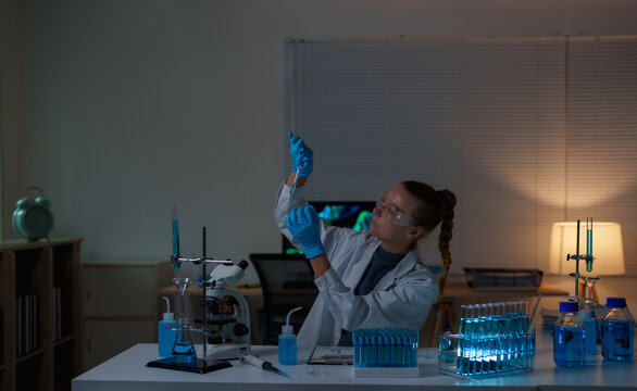Female scientist working in a research laboratory, pipetting blue liquid into a test tube for a scientific experiment, conducting medical diagnostics and chemical analysis late at night