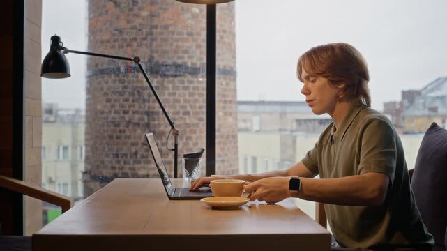 Caucasian young adult man drinking coffee while typing on laptop at cafe table. Perfect for advertising remote workflow, online learning, or digital nomad lifestyle