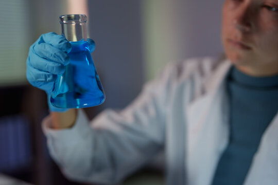 Scientist in a white coat wearing blue gloves inspects a conical flask of vivid blue solution during a focused laboratory experiment for chemical and medical research