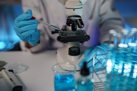 Scientist in protective gloves pipetting a blue solution onto a microscope slide, conducting research in a modern laboratory setting with various scientific equipment and test tubes