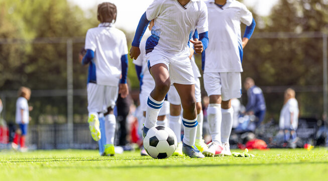 Junior Soccer Player Dribbling Ball on Pitch in White Kit - Youth Football Team Match Action and Ball Possession on Sunny Grass Field