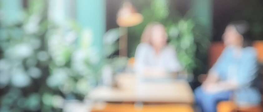 Blurred image of two people in a casual meeting in a cozy, green indoor setting with plants and warm lighting.