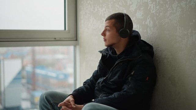 Close up of young man wearing headphones sitting by window with urban view hands clasped calm expression. Soft natural light illuminates scene background shows out of focus cityscape