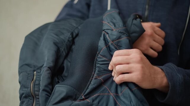Close up of man holding and examining interior lining of his blue jacket focusing on texture details like orange stitching. man wears dark clothing throughout with consistent hand