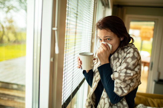 Sick woman with tissue and mug by window at home