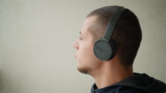 Close up of man wearing black over ear headphones facing sideways with minimal movement. subject has short dark hair and wears navy blue hoodie against plain light colored wall background