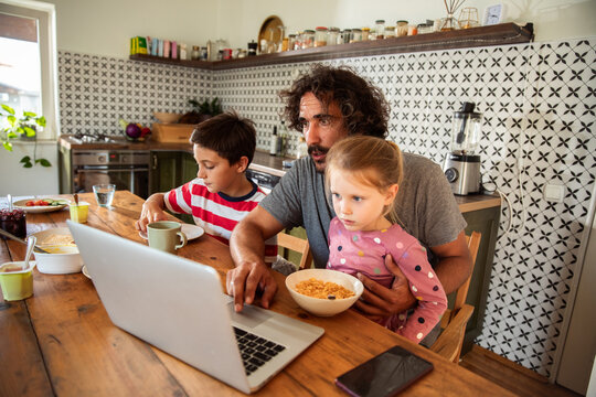 Father working on laptop with kids during breakfast in home kitchen