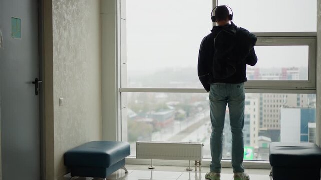 Man stands indoors near large window holding item and wearing headphones. setting includes modern interior elements like benches and radiator with cityscape visible through glass