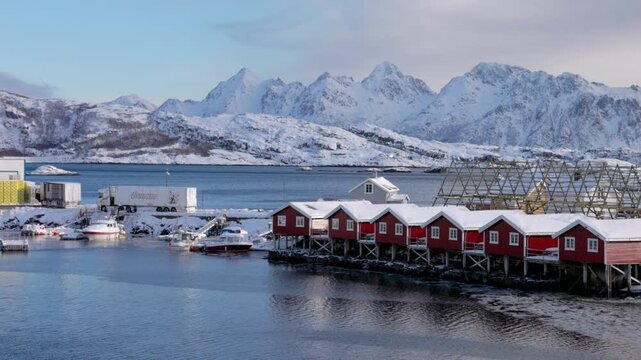 View of traditional red rorbuer cabins on stilts with fish drying racks against a backdrop of snow-covered mountains in Svolvaer, Nordland, Norway.