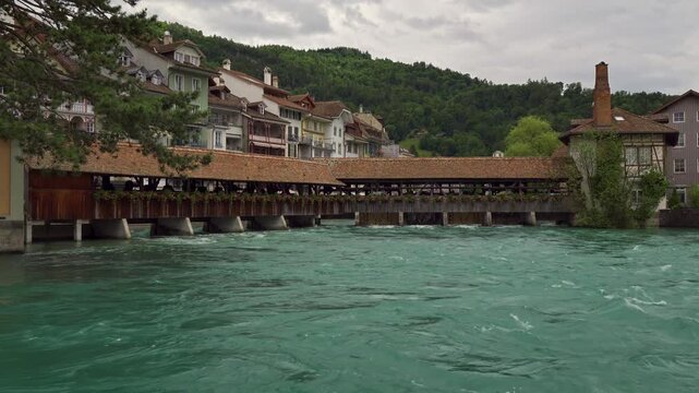 Turquoise Aare River flows beneath a flower-lined covered wooden bridge in Thun, Switzerland, with old town buildings and green hills. 4K UHD video.