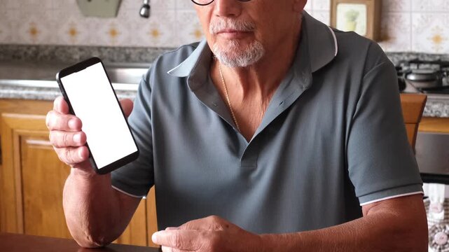 Elderly man with a goatee sitting at a kitchen table, using a lancet to prick his finger for a blood glucose test, and then showing a smartphone with a blank white screen to the camera