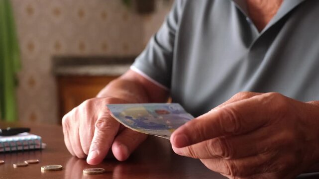 Close-up view of an elderly man's hands counting euro banknotes and coins on a wooden table, representing retirement savings, pension planning, and personal financial management at home