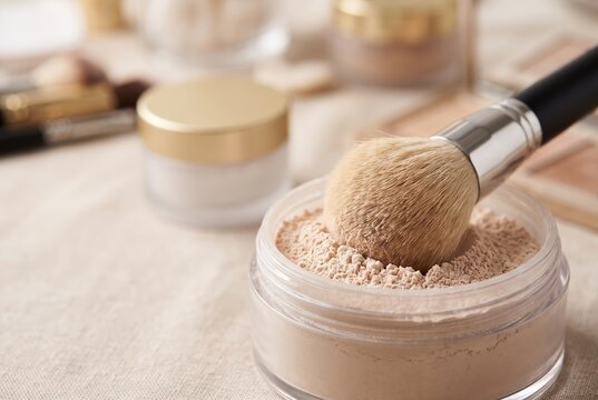 A makeup brush is dipped into a jar of loose powder on a table. Various cosmetics are nearby. The scene shows a beauty routine taking place, highlighting makeup products