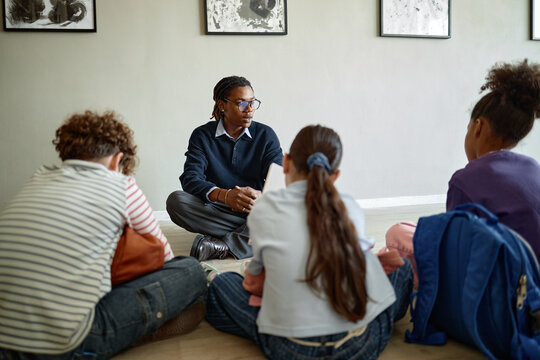 Black male museum guide talking to diverse group of children sitting on floor in art gallery. Image represents interactive learning and is ideal for educational marketing