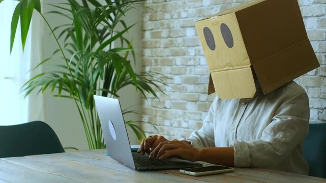 Unrecognizable person with a box on her head working remotely on a laptop computer, typing on the keyboard. Concept of employee alienation, modern work life, and online identity