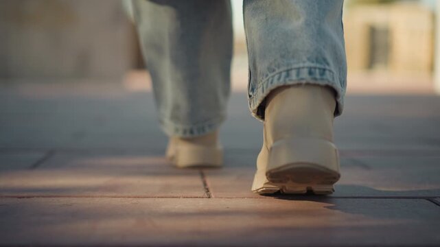Close up of person feet in light blue jeans and beige shoes walking steadily on sunlit pavement. focus remains consistently low angle capturing rhythmic movement from heel to toe with soft