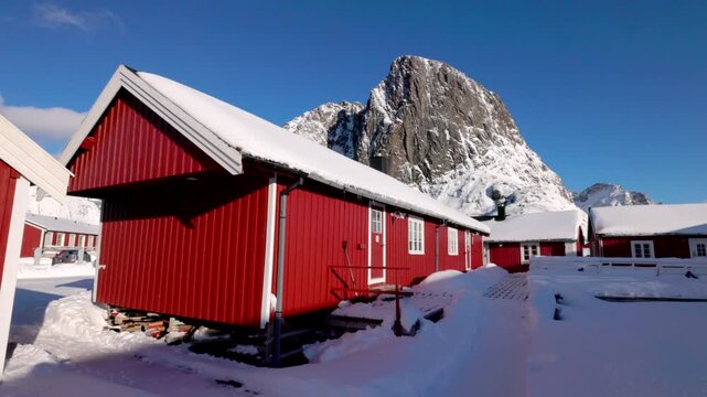 View of traditional red cabins covered in snow with mountains in the background in Hamnoy, Nordland, Norway.